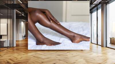 Close up of young dark skin woman with perfect body touching gently her hairless soft and silky legs after depilation in bed at home Wall mural